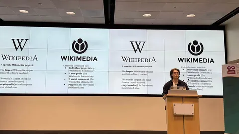 Chris Petrillo stands on stage in front of several large screens showing Wikimedia and Wikipedia logos