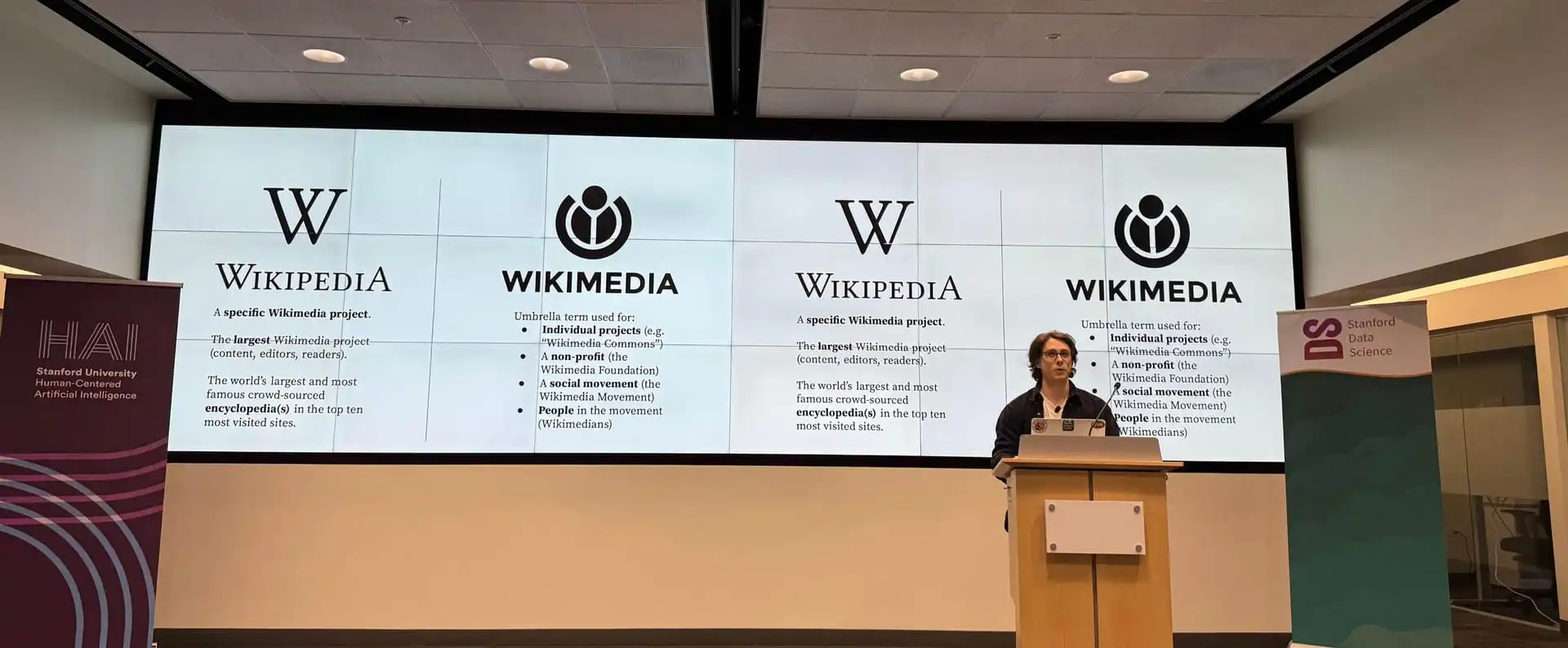 Chris Petrillo stands on stage in front of several large screens showing Wikimedia and Wikipedia logos