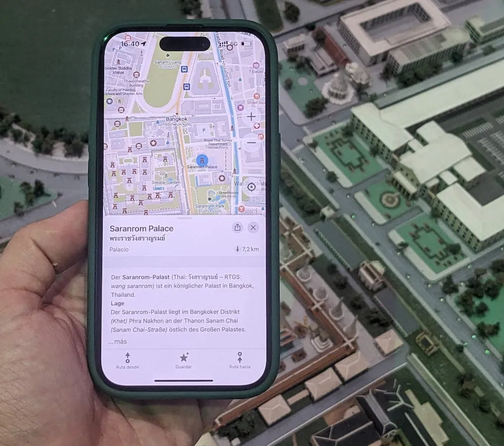 A hand coming in to the left side of the frame holding a smartphone with CoMaps open, showing information about Saranrom palace. Behind the phone is a miniature diorama of a city in Thailand.
