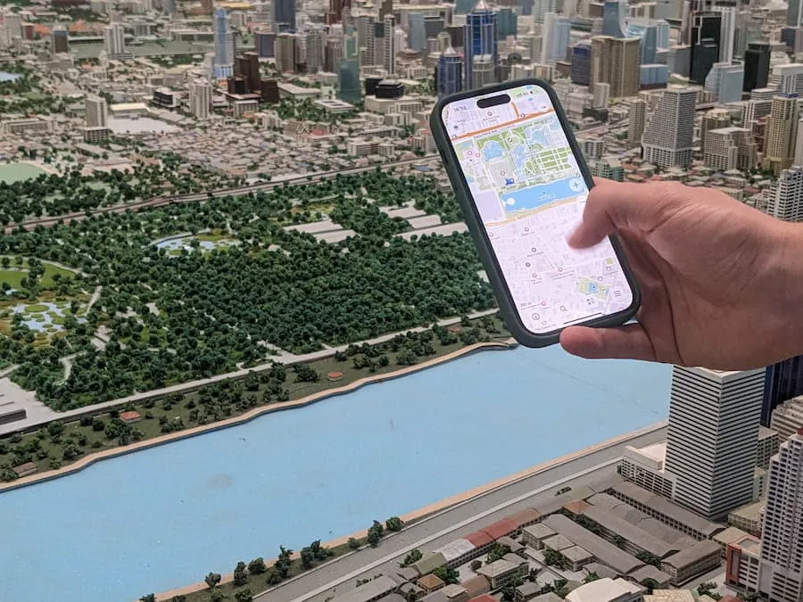 A hand coming in to the right side of the frame holding a smartphone with CoMaps open. Behind the phone is a miniature diorama of a city in Thailand.