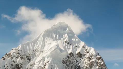 Photograph of the snow-covered summit of Mount Bradley against a blue sky, with a cloud drifting behind the peak.