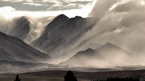 A photograph of an imposing landscape with jagged mountains in the background and a plain with trees in the foreground, everything is covered in a thick layer of cloud and mist swirling around the mountains.