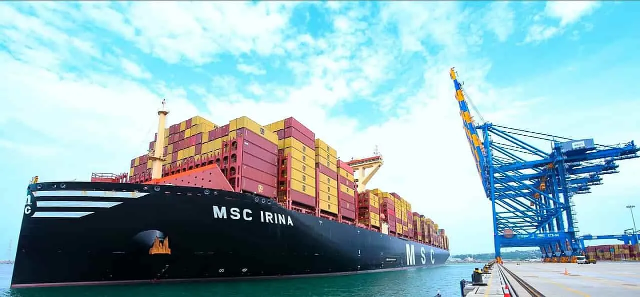 photograph of the largest container ship on earth, the MSC Irina, docked at a port with a blue cargo loading crane on the right.