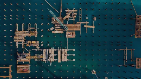 Aerial view of a pier under construction: a grid of pilings in teal water with cranes and platforms on top.