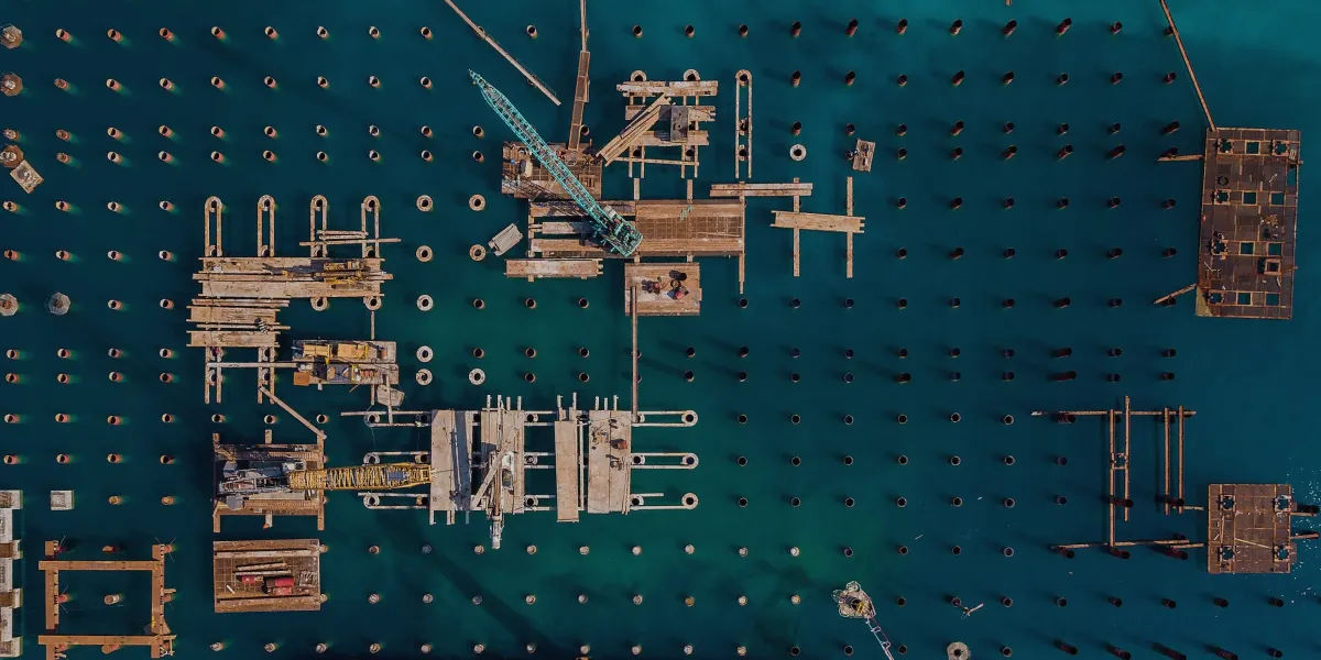 Aerial view of a pier under construction: a grid of pilings in teal water with cranes and platforms on top.