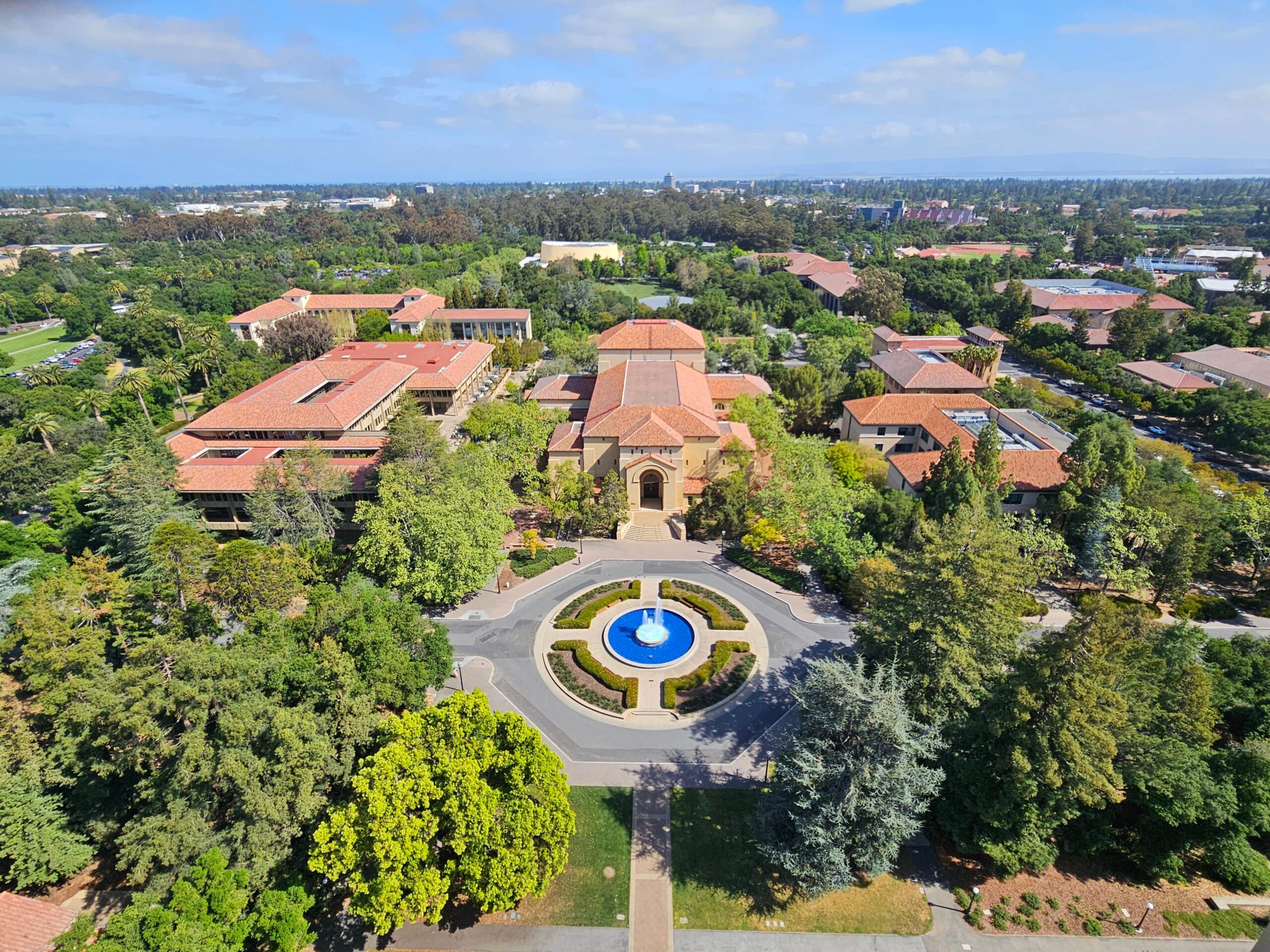 an aerial shot of the Stanford university campus, with many trees, a large square with a fountain in the centre, and different red roofed buildings.