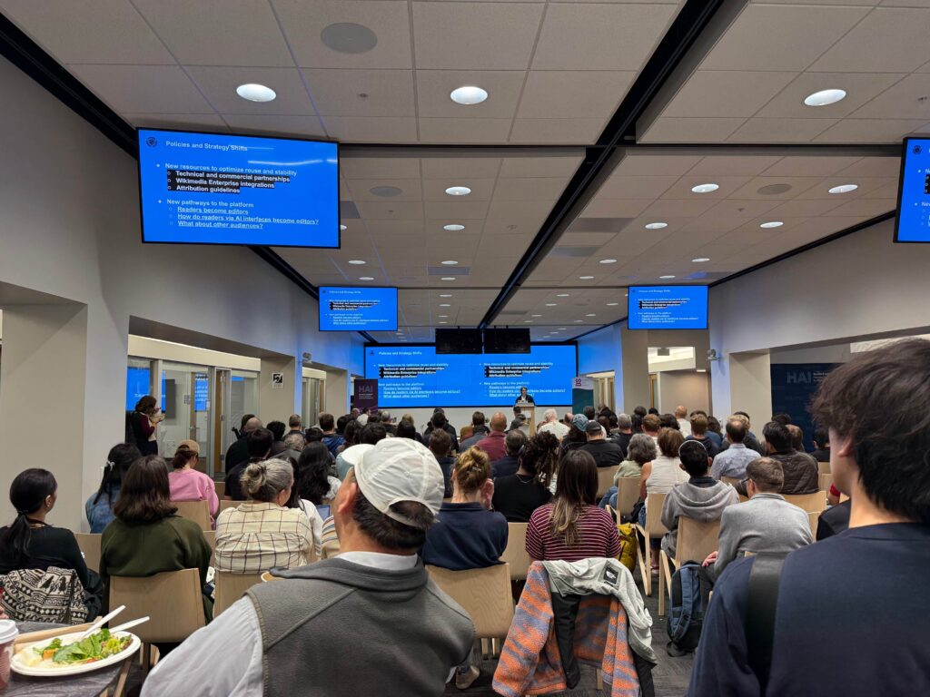 people sit on chairs looking at Chris Petrillo presenting at the Stanford lunch seminar, multiple screens showing a blue slides are visible overhead.