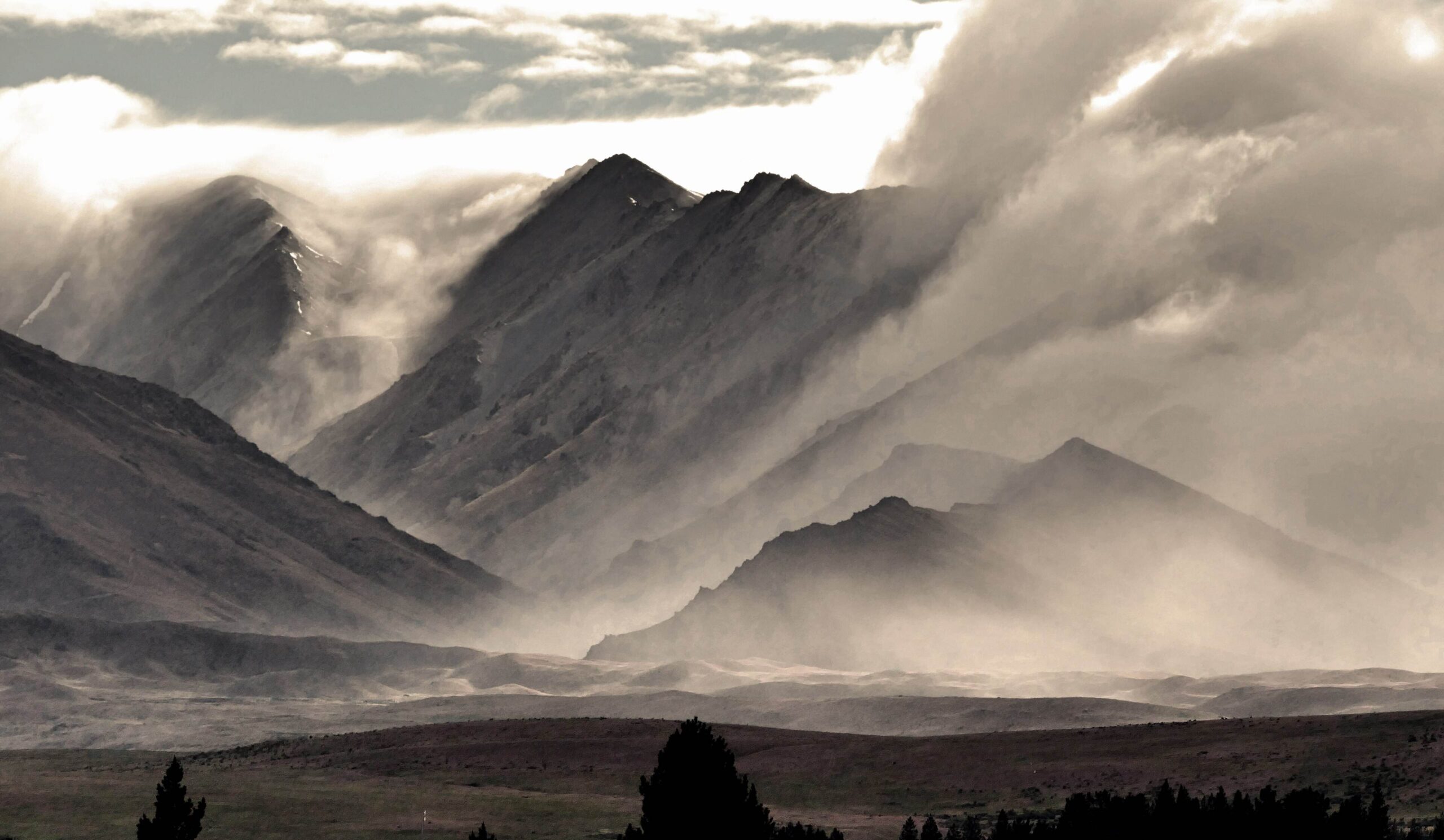 A photograph of an imposing landscape with jagged mountains in the background and a plain with trees in the foreground, everything is covered in a thick layer of cloud and mist swirling around the mountains.